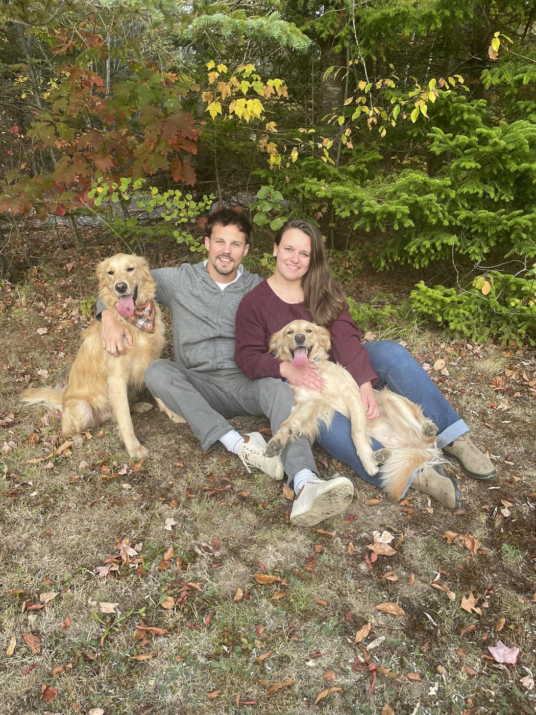 Micayla and her husband sitting in the woods with their two golden retrievers