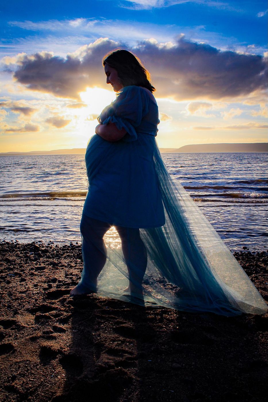 Expectant mother in a flowing blue gown on the beach at sunset