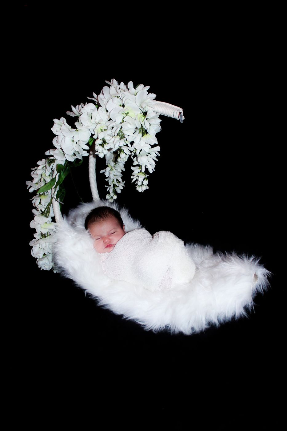 Newborn asleep on white fur beneath hanging white flowers